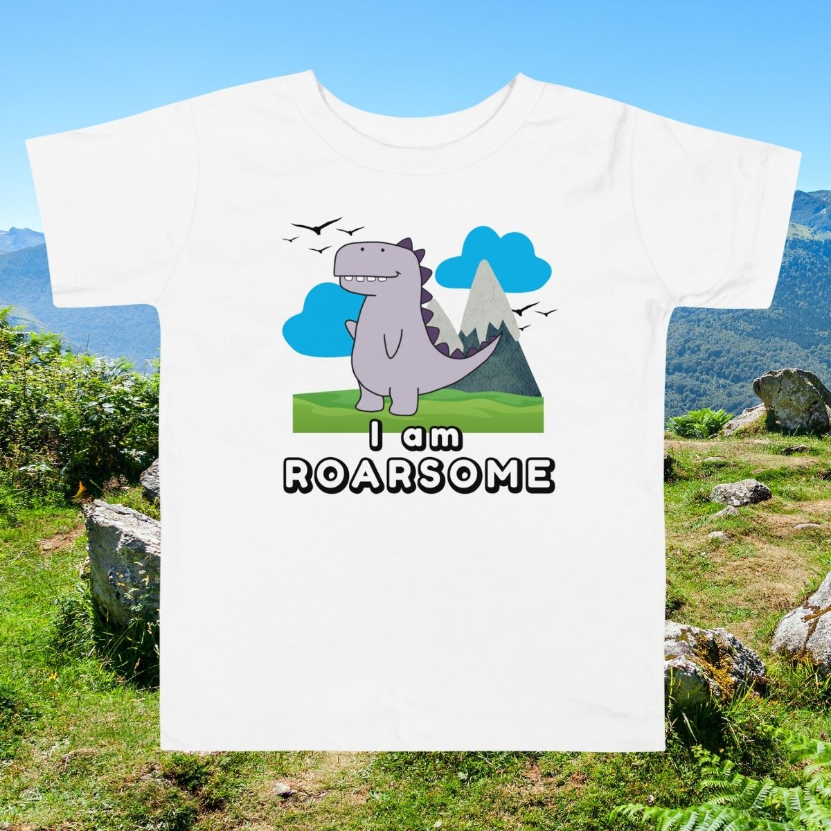 White toddler shirt with "I Am Roarsome" text and cartoon dinosaur in the center on a nature with greens and sky background—ideal for dino-loving kids.